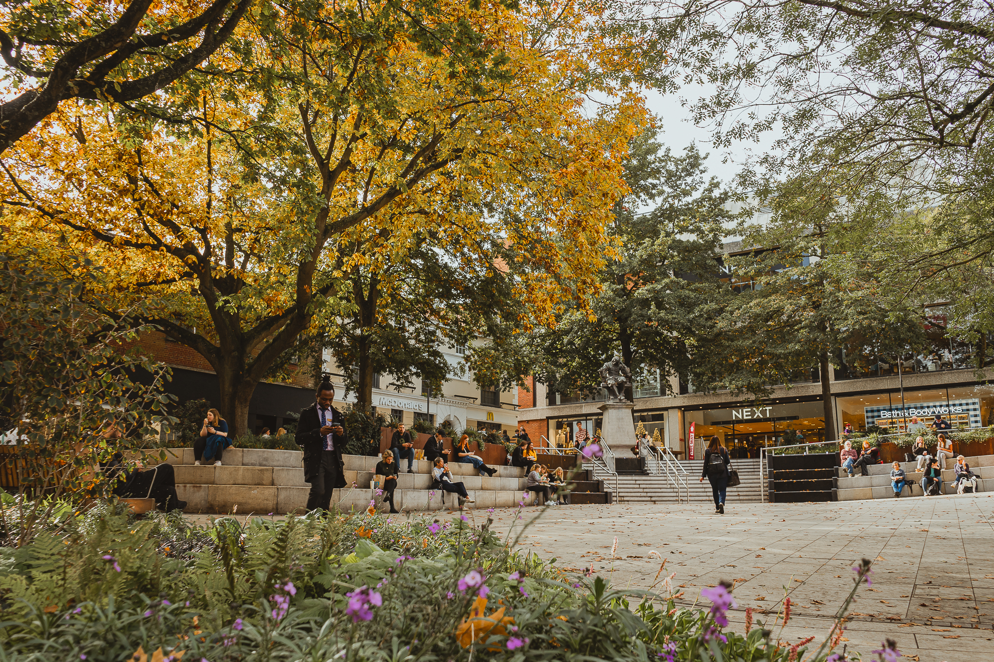 A view of the recently refurbished Hay Hill in autumn. In the foreground there are purple and orange flowers and greenery. A large open square is surrounded by stone steps, which people sit on underneath yellow-leafed trees. Entrance to the high street store NEXT is in the background.