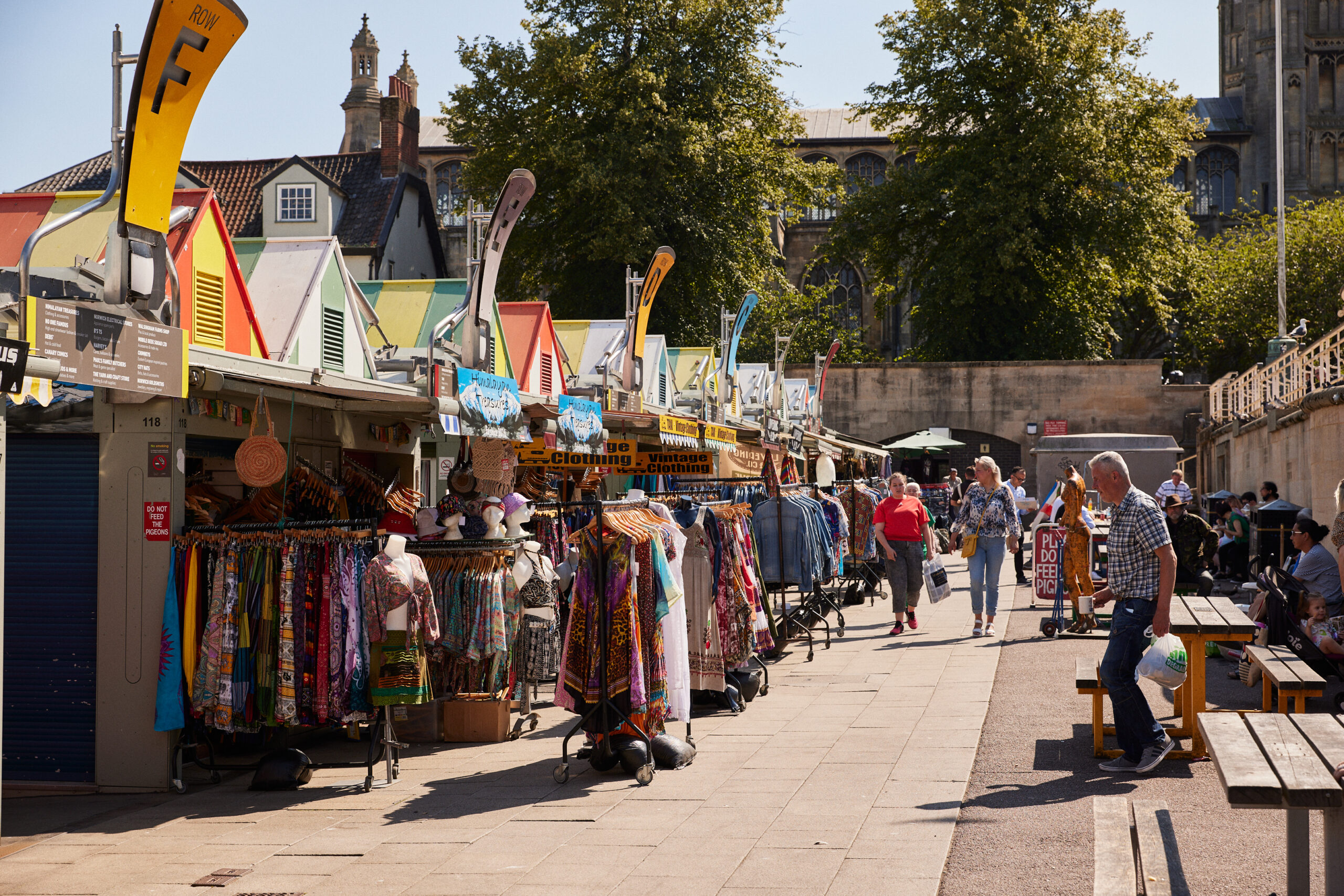 The back of Norwich Market in the sunshine. People sit at wooden benches along the right and walk between the benches and the colourful stalls on the left with rails of colourful clothes. There are leafy green trees in the background, where you can see the tops of buildings and St Peter Mancroft church.