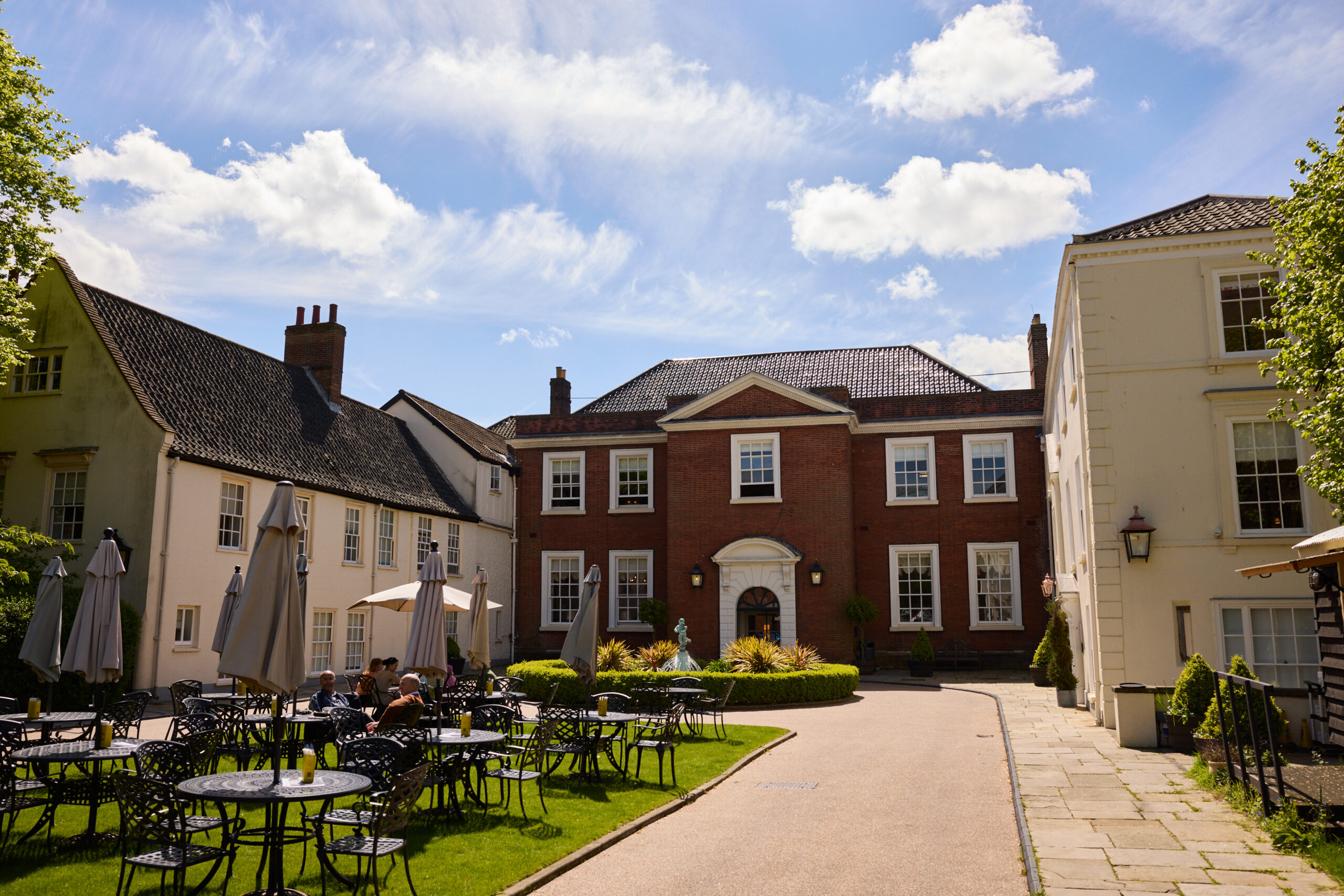 The frontage of The Assembly House in summer under a blue sky with white clouds. On the left the green lawn is covered with outside chairs, tables and parasols (some up, some down). The pathway leads to the front of the building which is brick with windows and door framed in white. The two wings on the left and right of the building are painted cream. In front of the entrance is a round hedge and fountain.
