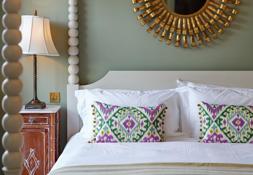 A close-up of the interior of on of The assembly House's bedrooms. On the left is a wooden cabinet with tall lamp with a white lampshade. The wall is sage green, with a gold round mirror. In front of the wall is a white four-poster bed (you can see two posts), with clean white sheets and pillows, layered with two brightly patterned purple and green cushions and a light green blanket.