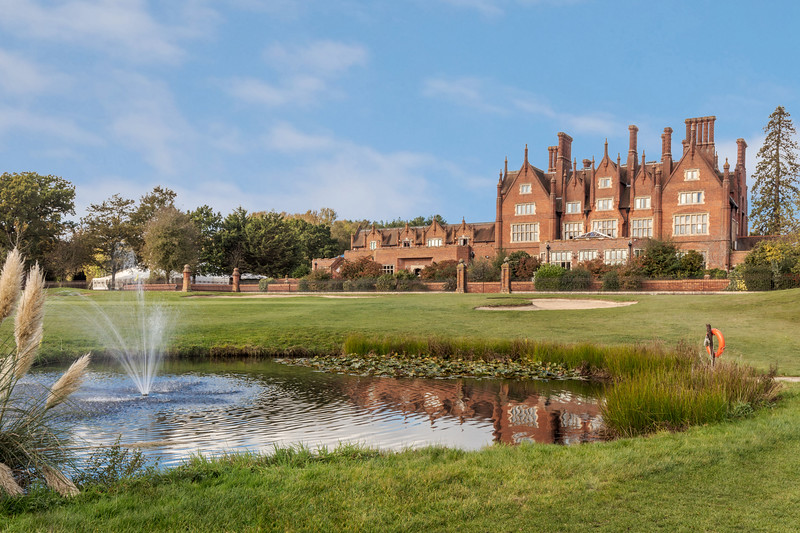 Dunston Hall sitting in it's manicured grounds with a lake and fountain at the forefront. There is a large lawn between the fountain and the hotel building, which is a red brick with multiple chimneys. On the left are trees.