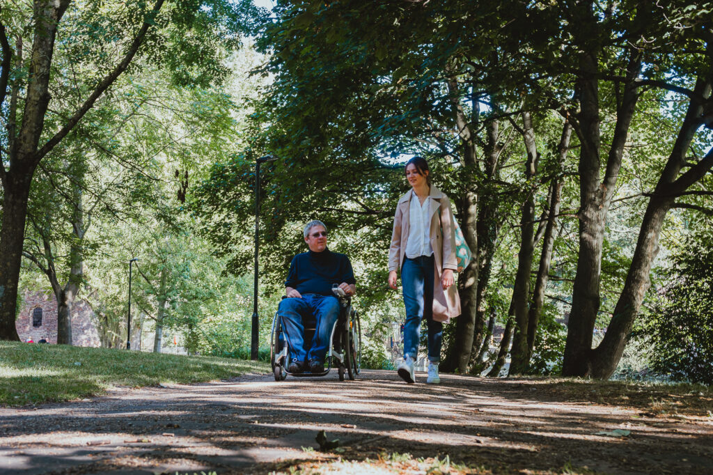 A man in jeans and a t-shirt in a wheelchair rolls alongside a walking woman in jeans, a shirt and pink jacket. They are surrounded by tall green trees and in the background, you can see a stone building on the left.